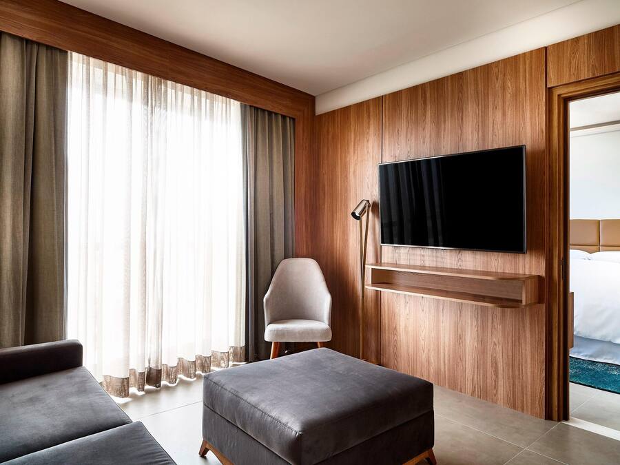 A hotel room with a large window, a flat-screen TV mounted on the wall, a grey sofa, a wooden panel wall, and a bed with white bedding.
