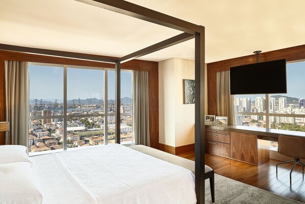Habitación de hotel con una cama grande, un escritorio con televisor y vistas a la ciudad a través de ventanales del suelo al techo.