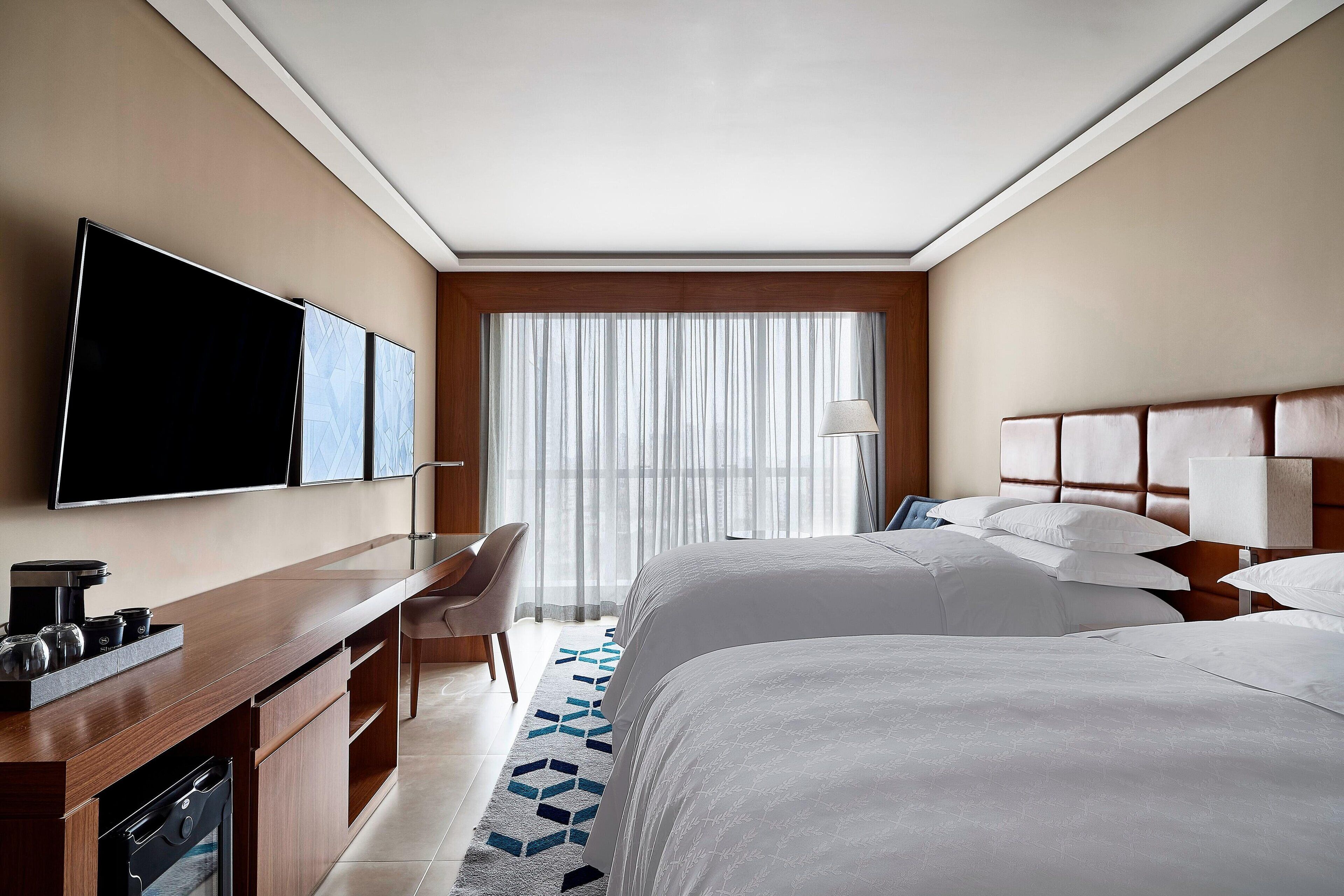 Habitación de hotel con televisión de pantalla plana, escritorio con cafetera, dos camas con cabeceros, una silla y un ventanal con cortinas translúcidas.