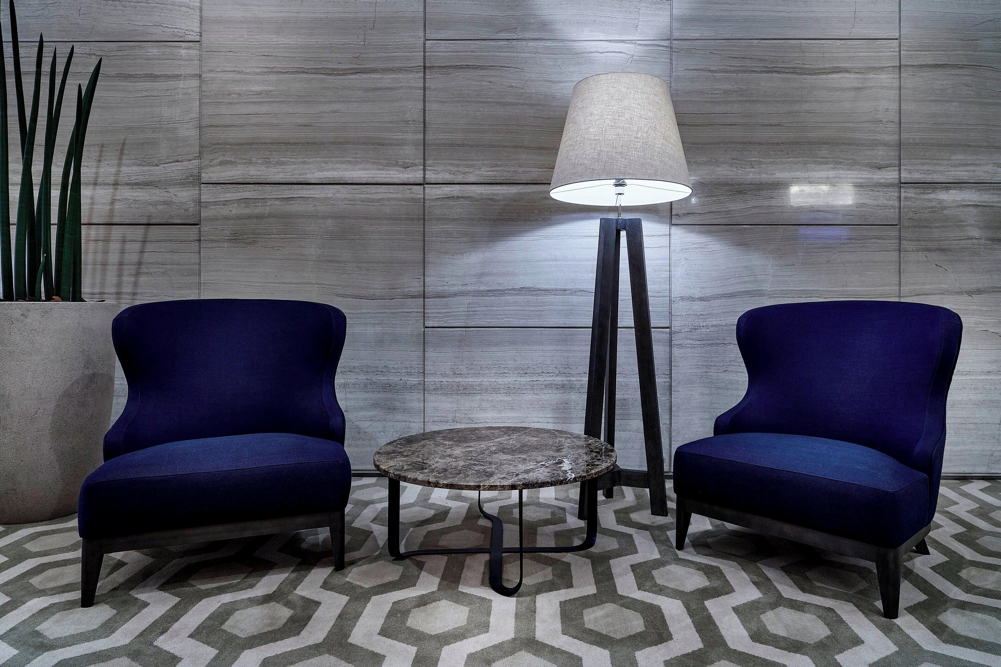 Two blue armchairs, a small round table, and a floor lamp in a room with a geometric patterned carpet and a grey tiled wall.