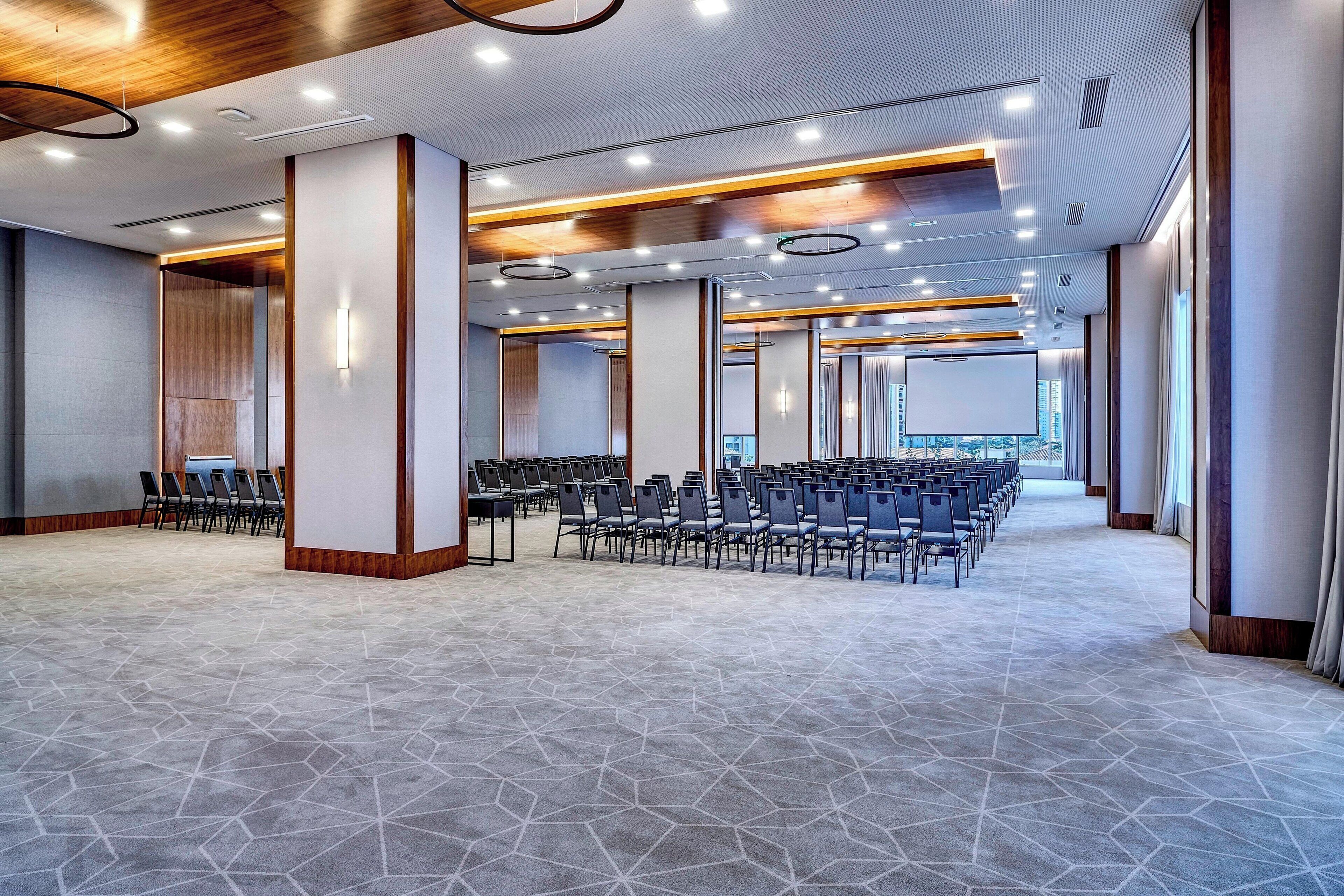 A spacious conference room with rows of chairs, a high ceiling with recessed lighting, and large windows allowing natural light.