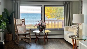 A room with a wooden rocking chair, a small round table with a flower vase, a white sofa, and a lamp. - Noc's Landing: Sun-Soaked Days & Starry Nights – Your Summer Escape Awaits! (Rapid River)