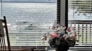 A room with a view of the sea through large windows, a round glass table with a vase of flowers, and two wooden chairs. - Noc's Landing: Sun-Soaked Days & Starry Nights – Your Summer Escape Awaits! (Rapid River)