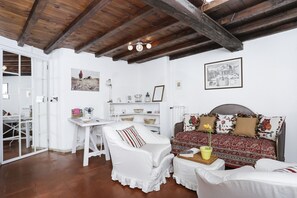 Une pièce avec un plafond en bois, un lit avec une literie à motifs, une chaise blanche, une petite table avec une plante et un bureau blanc avec des étagères.
