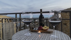 Deux verres à vin sont posés sur une table de balcon, accompagnés d’une bouteille de vin, d’une bougie et de quelques fruits.