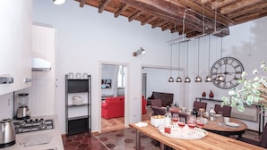 A modern kitchen with a gas stove, a coffee machine, and a sink. There is a dining area with a wooden table and chairs, and a large wall clock. - Regal Home in Trastevere 3BR (Roma)