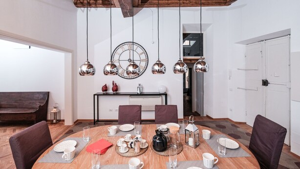 A dining area with a wooden table set for four, surrounded by chairs. A large wall clock and pendant lights hang above. - Regal Home in Trastevere 3BR (Roma)