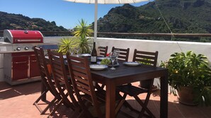 An outdoor patio with a table, chairs, a grill, and a view of a mountainous landscape.