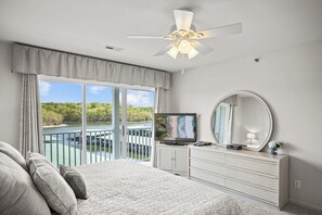 Une chambre dotée d’un grand lit, d’un ventilateur de plafond, d’une télévision, d’une commode, d’un miroir et d’un balcon offrant une vue sur un lac et des arbres.