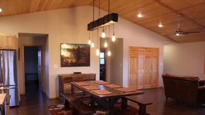 A spacious living area with a wooden dining table, benches, and a red patterned rug. There is a wooden cabinet, a painting on the wall, and a ceiling fan. - Blacktail Inn & Stations (Dillon)