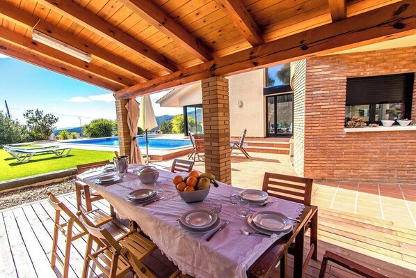 Un espace extérieur abrité, aménagé pour les repas, avec vue sur la piscine, doté de mobilier en bois et d’un mur en briques.