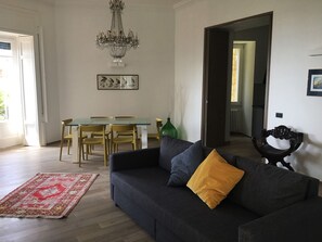 A living room with a dark grey sofa, a yellow pillow, a red and white rug, a chandelier, a dining table with chairs, and a framed artwork on the wall. - Casa del Cedro superior apartment with private garden on the Gulf of Naples. (Napoli)