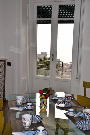 A dining area with a glass table set for four, a vase with flowers, and a city view through a door with shutters.