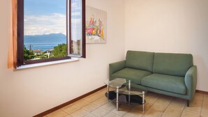 A room with a green sofa, a glass table, a painting, and a window with a view of mountains and a body of water. - Apartment Rosa Portland With Pool (Lazise)
