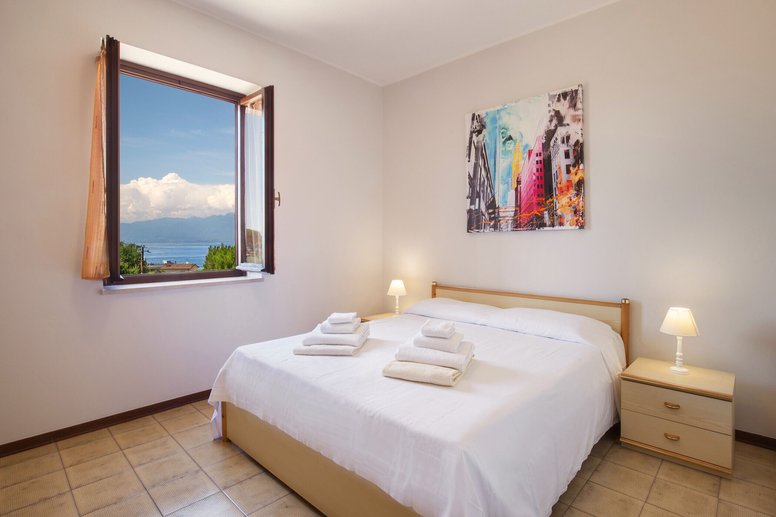 A bedroom with a bed, two bedside tables, a window with a view, and a painting on the wall.