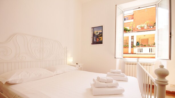 A hotel room with a bed, white bedding, a nightstand, a wall-mounted painting, and an open window with a view of the outside balcony. - Walk to the Colosseum from your Family Apt (Roma)