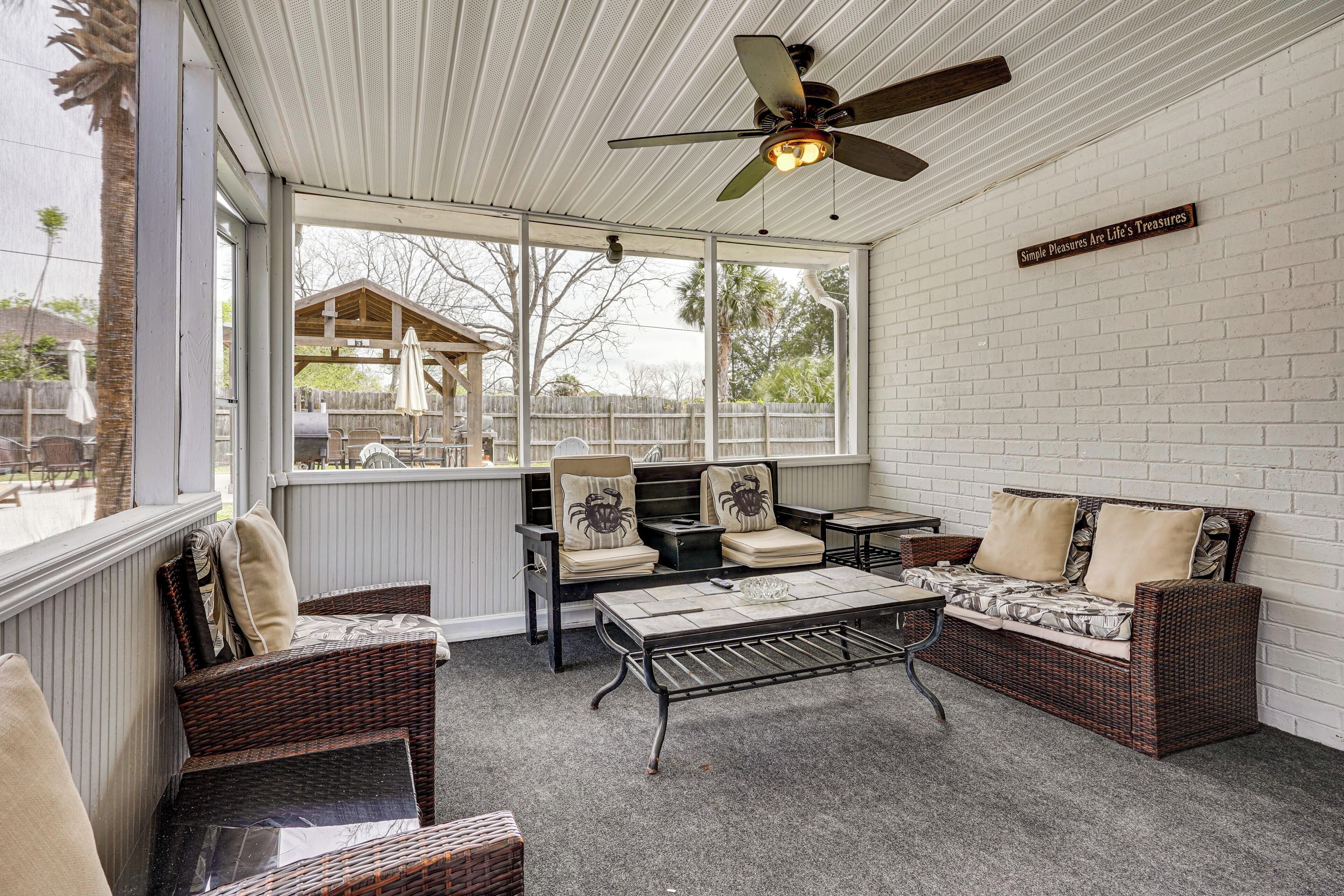 A covered patio with wicker furniture, a ceiling fan, and a view of a wooden structure in the background.