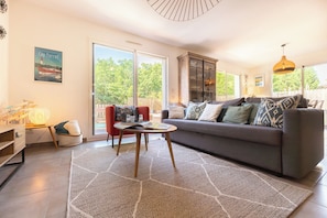 A modern living room with a grey sofa, a red armchair, a coffee table, and a large window offering a view of greenery.