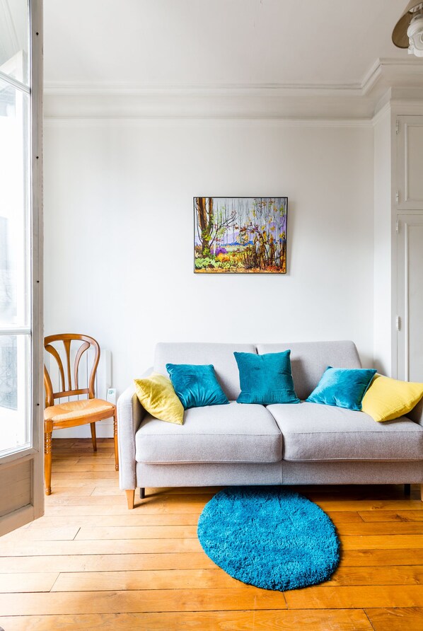 Un salon avec un canapé gris, des coussins colorés, un tapis bleu et un tableau accroché au mur.