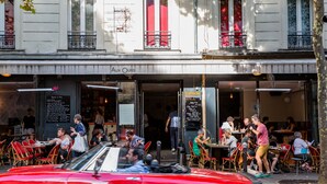 A restaurant with outdoor seating, a red car parked in front, and a building with balconies.