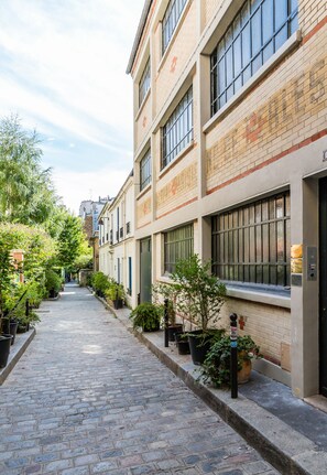 Une ruelle étroite pavée, bordée d’immeubles aux grandes fenêtres et agrémentée de plantes en pot.