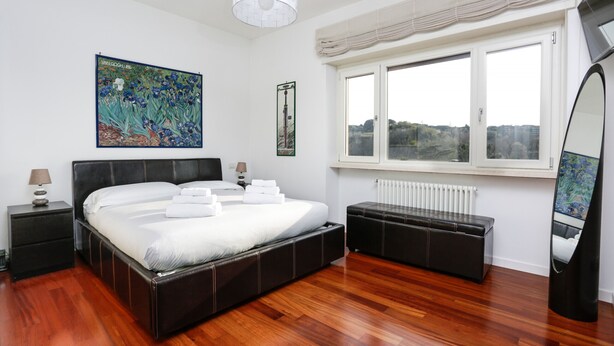 A modern bedroom with a large bed, a bedside table, a bench, a window with a view, and a mirror. - Enjoy the Sunset on Your Vatican Terrace (Roma)