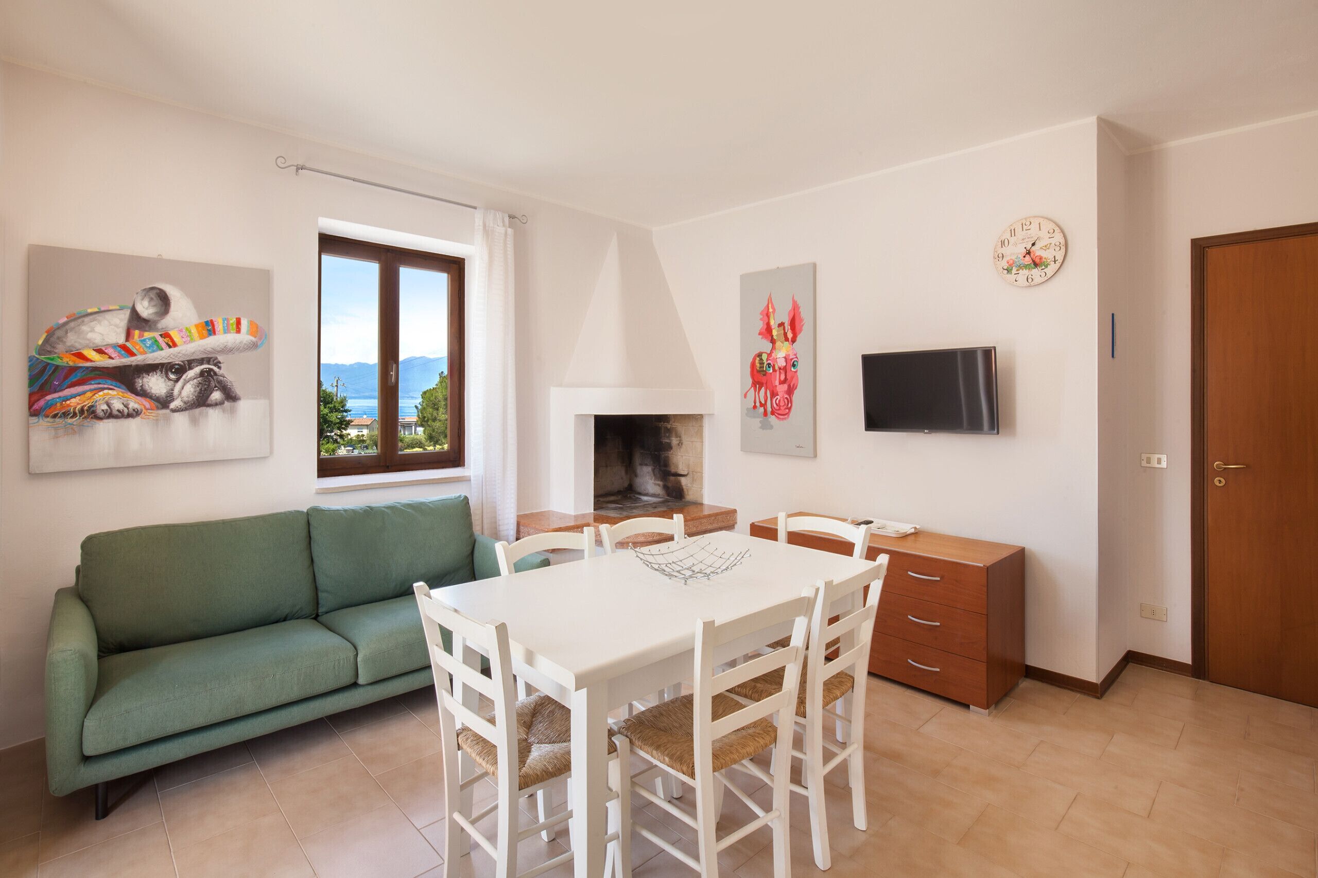 Een woonkamer met een groene bank, een witte eettafel met stoelen, een open haard, een televisie en kunstwerken aan de wanden.