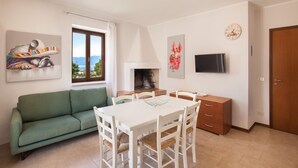 A living room with a green sofa, white dining table, and chairs, a fireplace, a TV, and artwork on the walls. - Apartment Rosa Sweet Sara With Pool (Lazise)