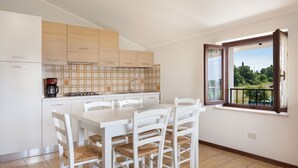 A modern kitchen with white cabinets, a dining table with white chairs, and a window with a view of greenery. - Apartment Rosa Noisette With Pool (Lazise)
