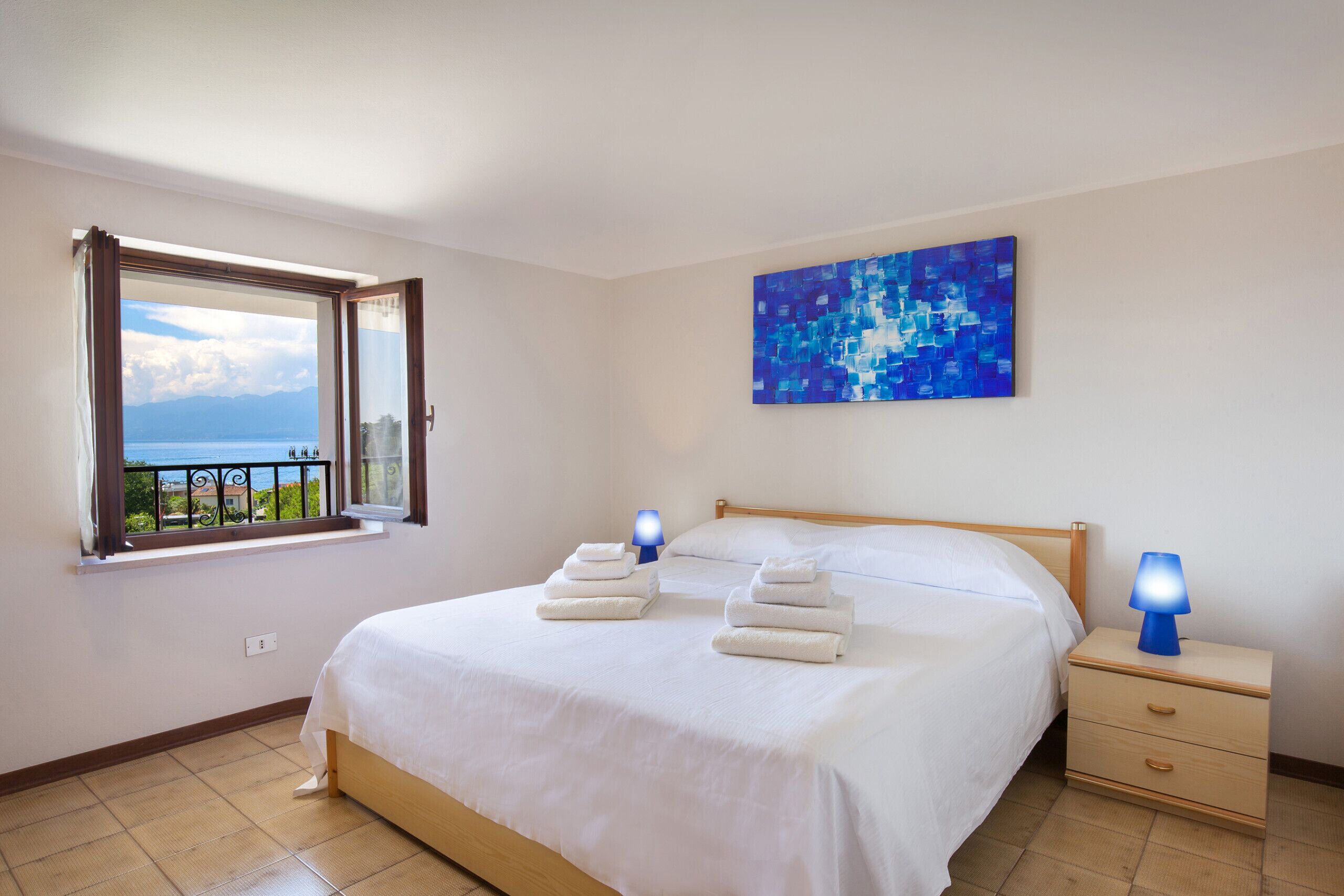 Een net opgemaakt bed met witte lakens, een blauw abstract schilderij aan de muur, een nachtkastje met een lamp, en een raam met uitzicht op de zee.