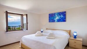 A neatly made bed with white linens, a blue abstract painting on the wall, a nightstand with a lamp, and a window with a view of the sea. - Apartment Rosa Noisette With Pool (Lazise)