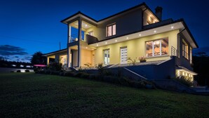 A two-story house with a balcony, large windows, and a well-lit exterior at dusk.