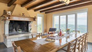 A dining area with a large wooden table, chairs, a fireplace, and a view through large windows.
