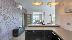 A modern kitchen with a marble backsplash, black cabinets, and a central island. There are pendant lights above the island and a view into the living area through an open door.