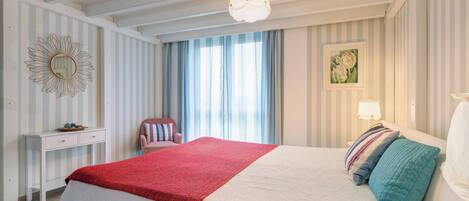Un dormitorio con cama, mesita de noche, silla, ventana con cortinas, un reloj de pared y un cuadro.