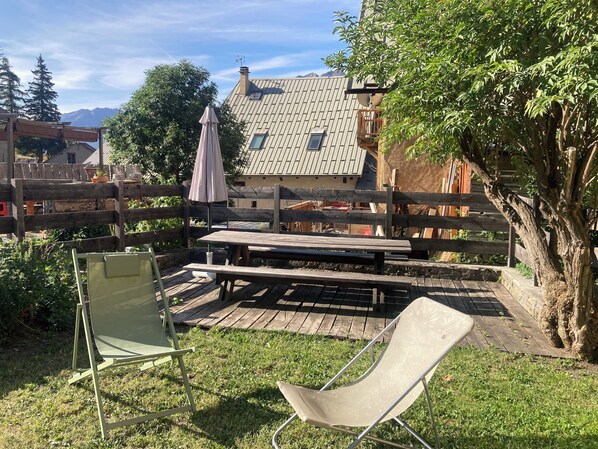 Um deck de madeira com uma mesa de piquenique e cadeiras, cercado por um gramado e uma árvore.