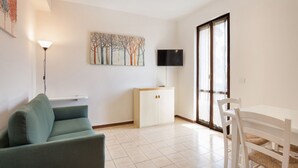 A living room with a green sofa, a white cabinet, a dining table with white chairs, a flat-screen TV mounted on the wall, and a large window with blinds. - Apartment Rosa Bourbon With Pool (Lazise)