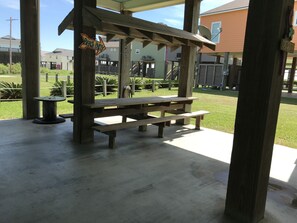 Ein Holztisch mit Bänken unter einem Schutzdach, umgeben von einer Rasenfläche und Häusern im Hintergrund.