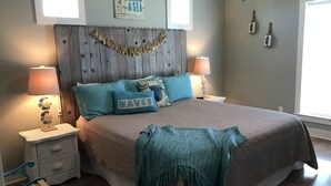 Un dormitorio con cabecera de madera, dos mesitas de noche con lámparas, una cama con manta y almohadas color turquesa, y una pared con un cartel que dice 'SAND & SEA'.