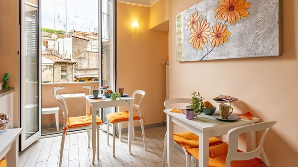 Un comedor con sillas y mesas blancas, un cuadro de flores en la pared y vistas de edificios a través de un ventanal.