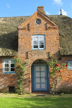 Une maison en briques avec un toit de chaume, une porte bleue et un petit jardin parsemé de fleurs rouges.