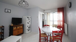 A dining area with a white table and red chairs, a black refrigerator with decorative magnets, a TV on a wooden cabinet, and a geometric pendant light. - 2 room apartment with sea view and parking (Canet-en-Roussillon)