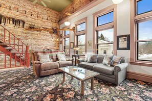 A cozy living room with a brick wall, large windows, and a staircase. - Historic Hardware Loft | 2 Blocks to Downtown (Saugatuck)