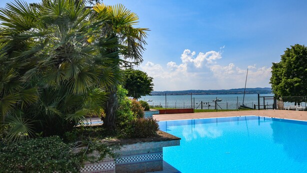 En swimmingpool med udsigt til havet, omgivet af frodig vegetation og en fliseterrasse.