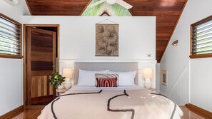 Quarto com teto de madeira, uma cama grande com manta bege e vermelha, duas luminárias de cabeceira e uma obra de arte na parede.