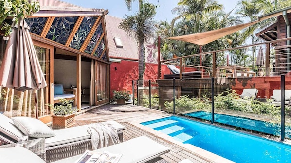 Uma área à beira da piscina com um deck de madeira, cadeiras de descanso e um salão envidraçado com vista para a piscina.