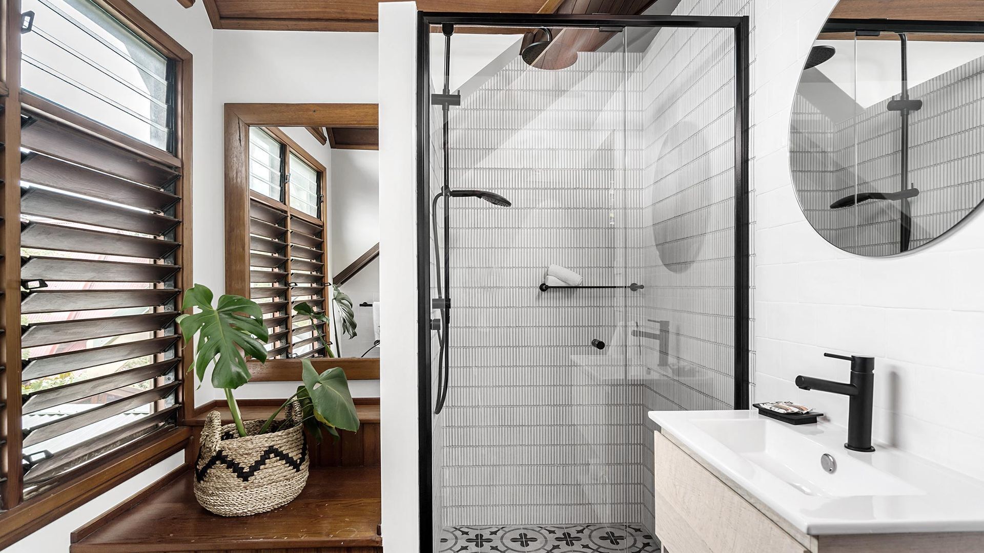 Un baño moderno con ducha, lavabo y espejo. Hay una planta en el suelo y una ventana con persianas.