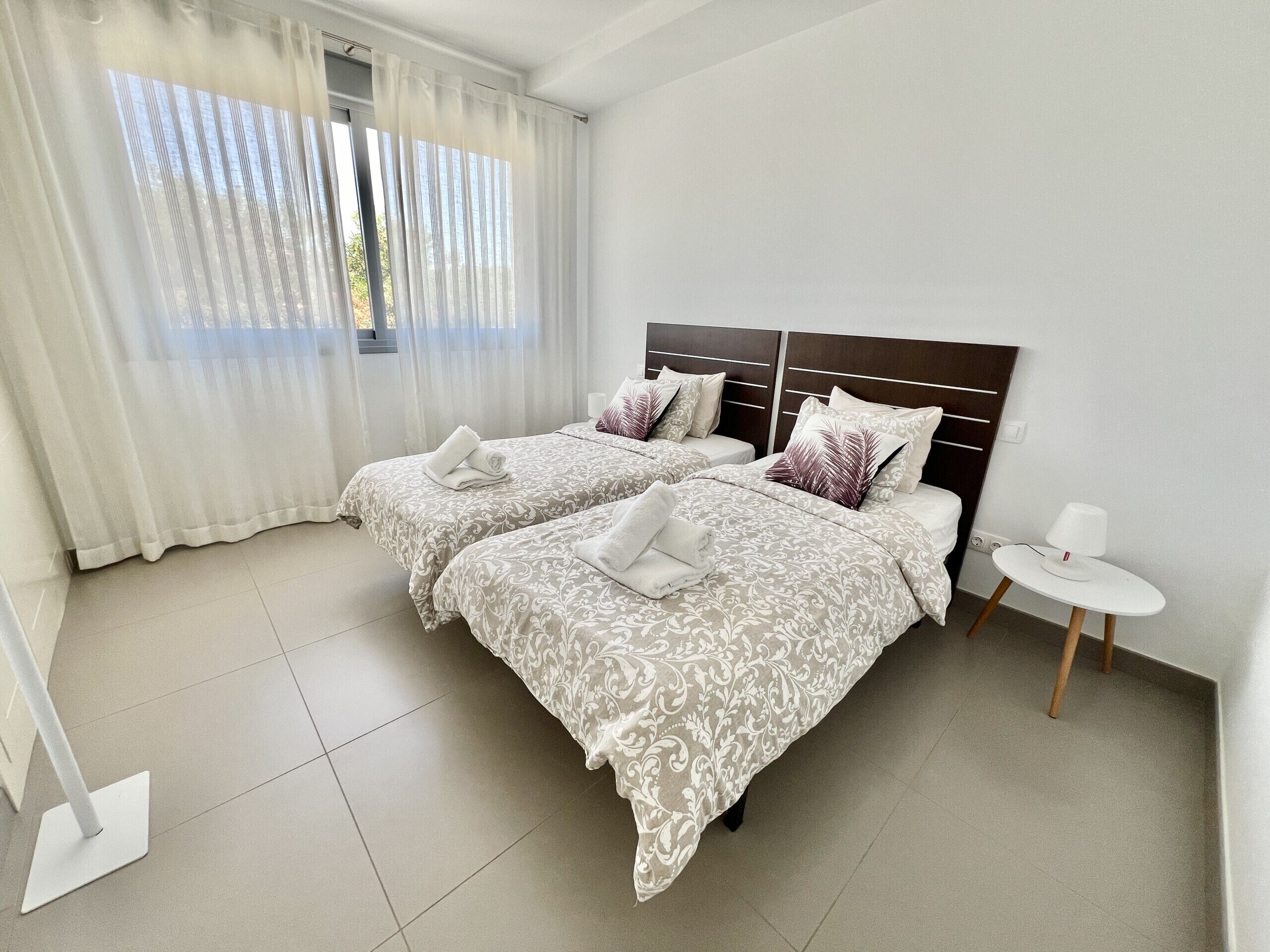 Une chambre d’hôtel avec deux lits, une petite table blanche et une fenêtre avec des rideaux légers.