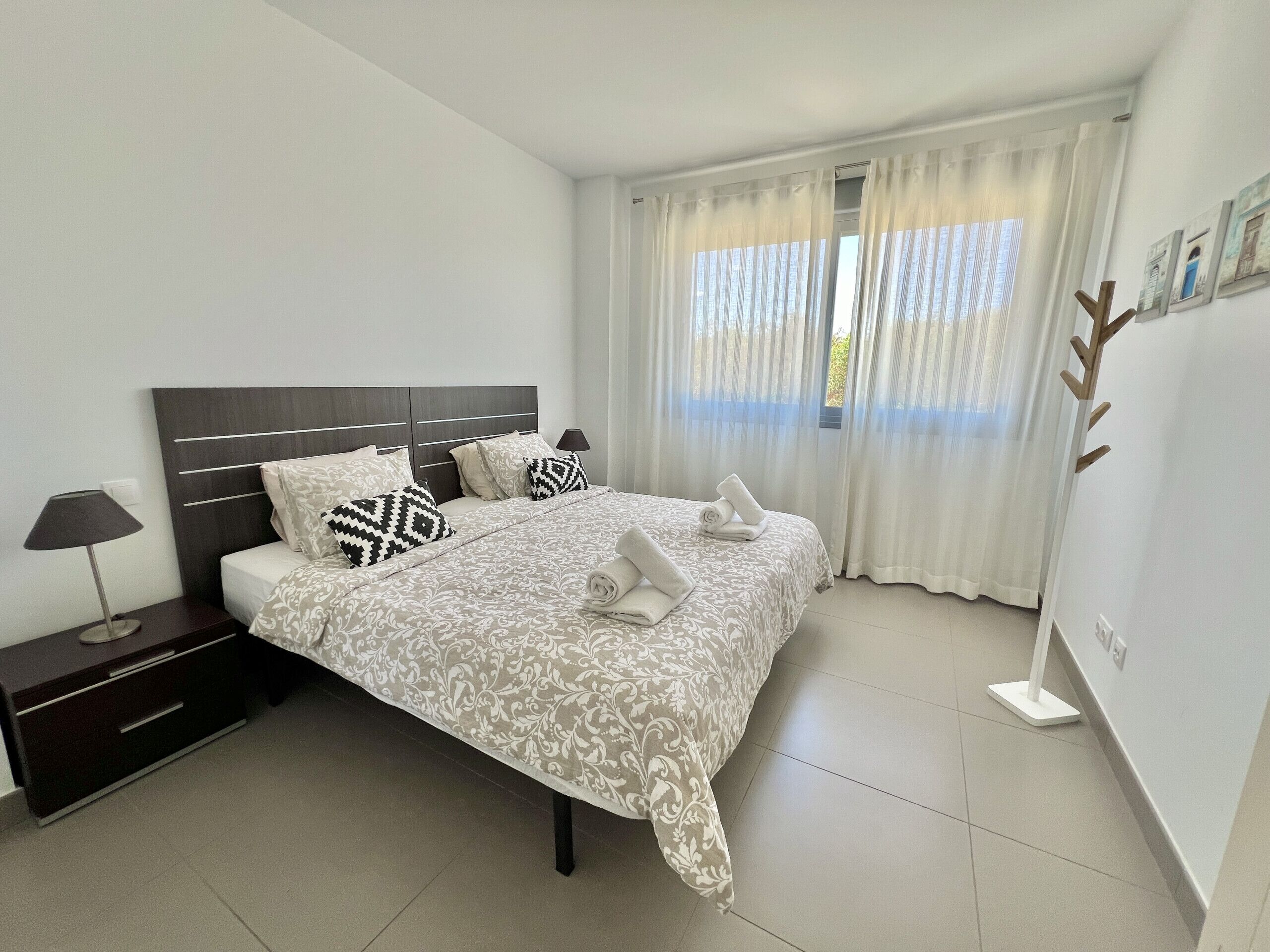 A neatly made bed with patterned bedding, a bedside table with a lamp, and a window with sheer curtains.