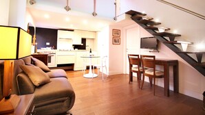 A modern living room with a sofa, a dining table with chairs, a staircase, and a kitchen in the background.